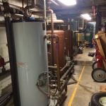 A large water heater in a warehouse surrounded by pipes and various equipment, showcasing an industrial setting.